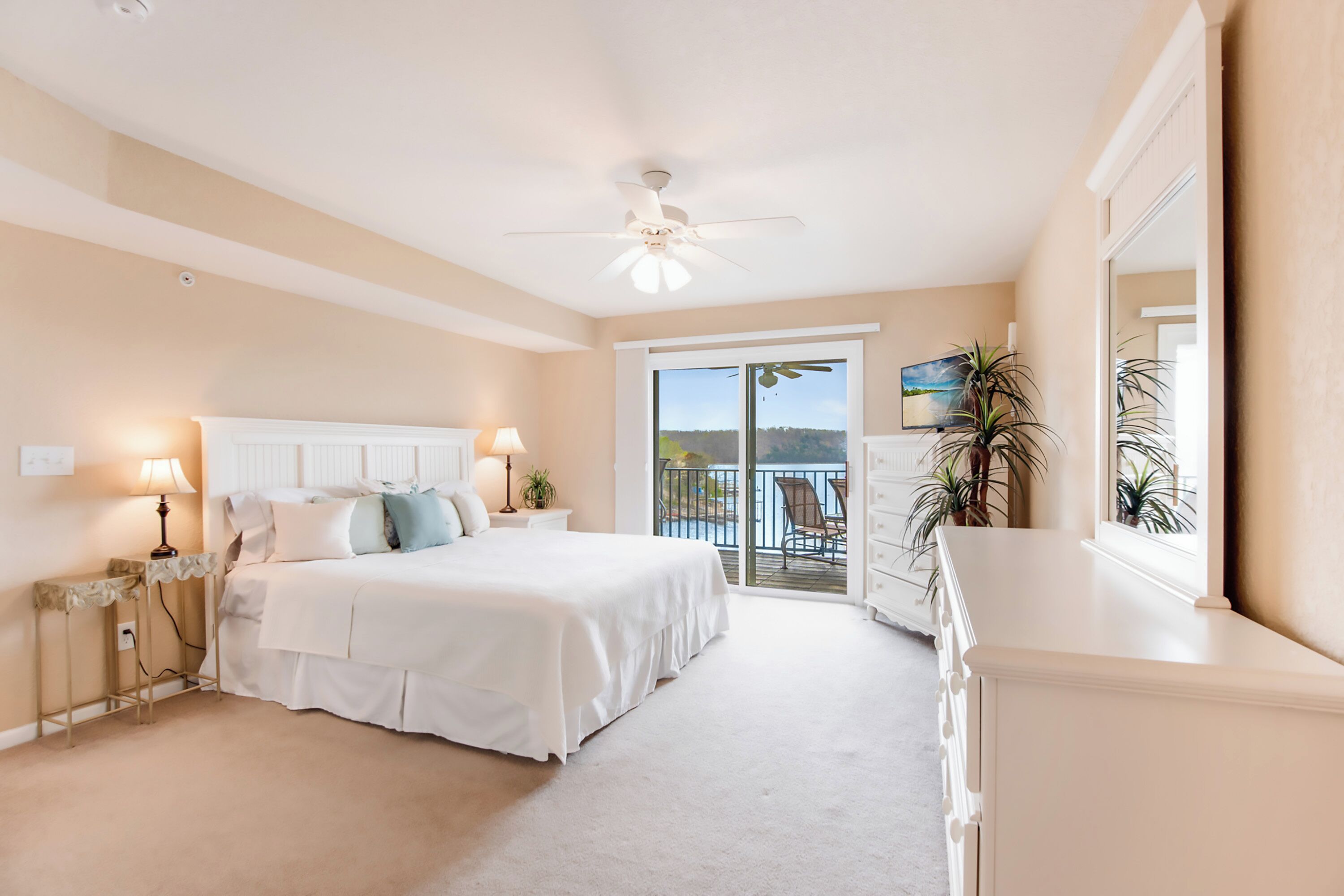 A bedroom with a bed, bedside lamps, a ceiling fan, a balcony with a view, and a dresser.