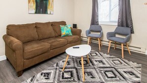 Een woonkamer met een bruine bank, twee grijze stoelen, een klein wit tafeltje en een vloerkleed met een patroon.