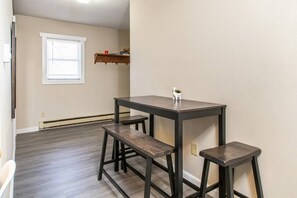 Ein Essbereich mit Tisch und zwei Bänken, eine kleine Regalanlage mit Büchern sowie ein Fenster mit Jalousien.