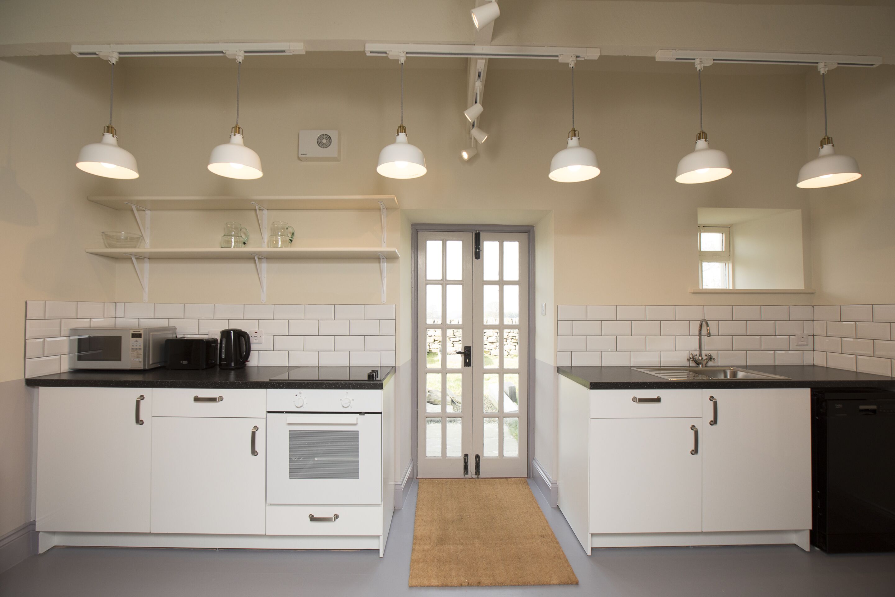 Une cuisine avec des meubles blancs, un four à micro-ondes, une machine à café et un évier. Il y a des luminaires au plafond et une fenêtre.