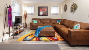 Ein Wohnzimmer mit einem braunen Ecksofa, einem Flachbildfernseher auf einem Standfuß, einem farbenfrohen Teppich und abstrakten Wandbildern.