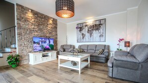 Una sala de estar con una pared de piedra, un televisor de pantalla plana, un sofá gris, una mesa de centro blanca y una escalera.