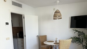 Ett modernt vardagsrum med en matplats, ett kök i bakgrunden, en platt-TV som är monterad på väggen och en krukväxt.