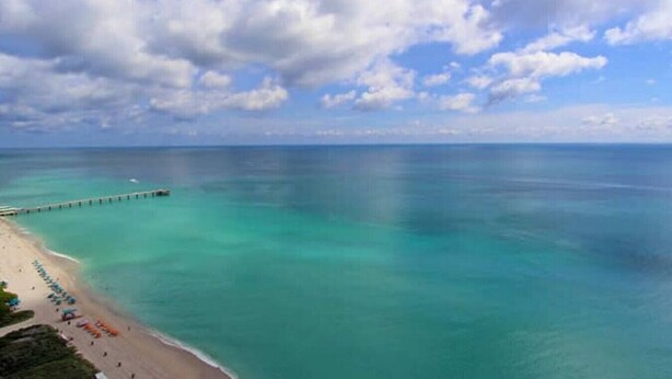 A beach with clear turquoise water, a pier extending into the sea, and a line of umbrellas on the sand.