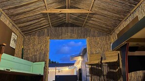A room with a thatched ceiling, wicker furniture, and a view of the outside at dusk.