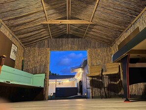 A room with a thatched ceiling, wicker furniture, and a view of the outside at dusk.
