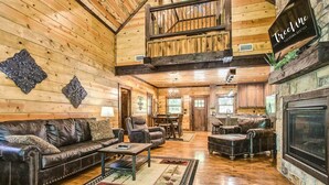 A spacious living room with a leather sofa, a coffee table, and a fireplace. There is a dining area with a table and chairs, and a staircase leading to an upper level. - Woodland Hot Tub Retreat for Families and Friends (Broken Bow)