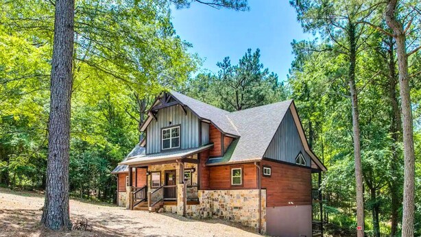 A two-story log cabin with a stone foundation, a front porch, and a steeply pitched roof, surrounded by a forest. - Woodland Hot Tub Retreat for Families and Friends (Broken Bow)