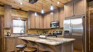 A modern kitchen with wooden cabinets, a stainless steel refrigerator, a granite countertop, and pendant lighting. - Woodland Hot Tub Retreat for Families and Friends (Broken Bow)