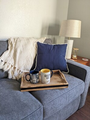 A grey sofa with a white knitted throw, a blue pillow, and a tray with two mugs. - Historic 2-bedroom Sam Hughes Casita w/ Courtyard (Tucson)