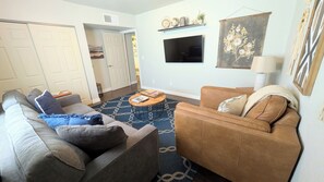 A living room with a sofa, a coffee table, and a TV. There is a door leading to another room and a wall-mounted shelf with decorative items. - Historic 2-bedroom Sam Hughes Casita w/ Courtyard (Tucson)