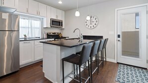 A modern kitchen with white cabinets, stainless steel appliances, a central island with bar stools, a microwave, and a wall clock.