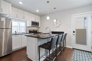 A modern kitchen with white cabinets, stainless steel appliances, a central island with bar stools, a microwave, and a wall clock. - Historic 2-bedroom Sam Hughes Casita w/ Courtyard (Tucson)