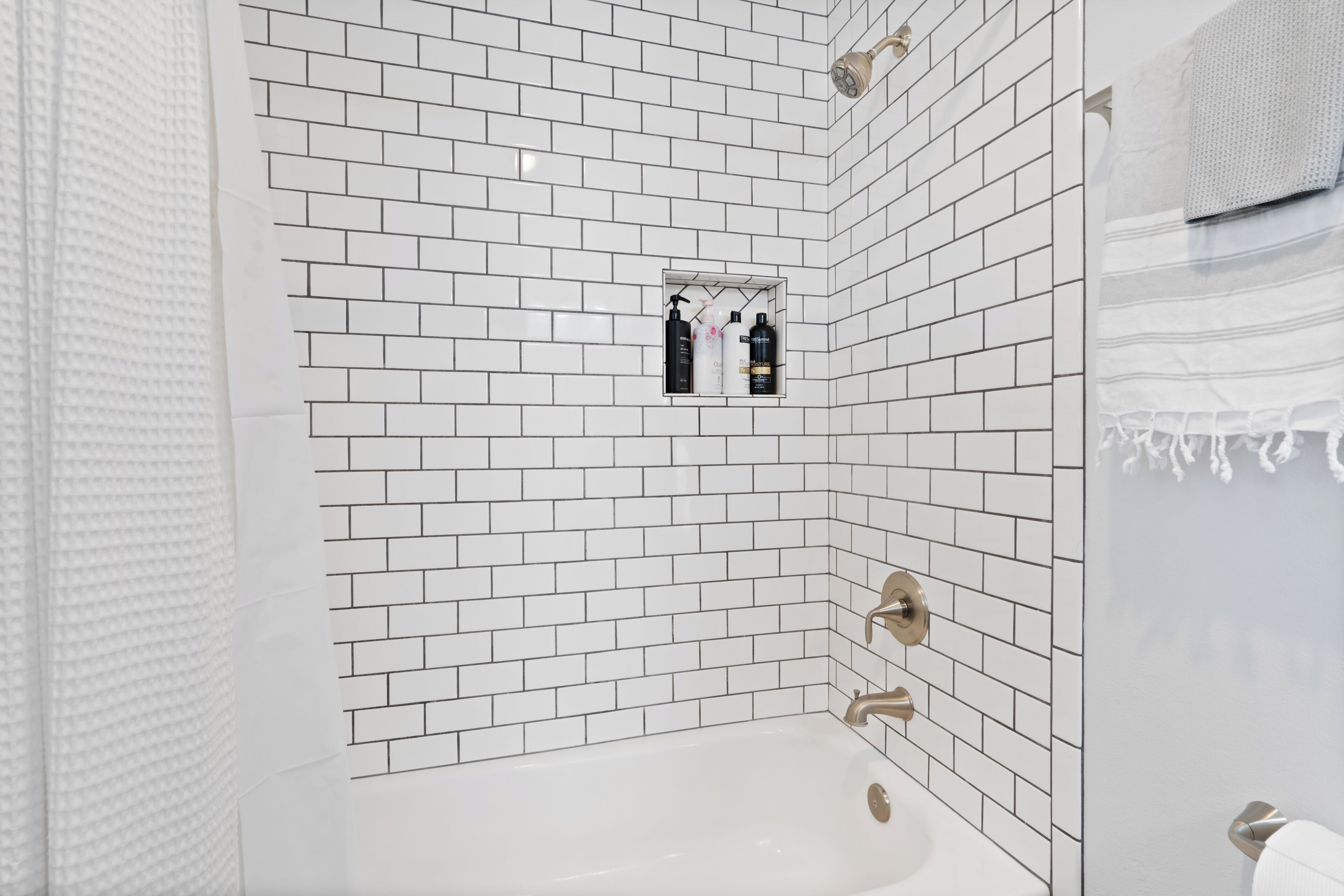 Une salle de bain avec des carreaux de céramique blancs rectangulaires, une baignoire et une étagère contenant des articles de toilette.