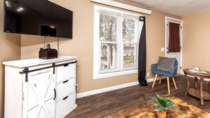A room with a flat-screen TV mounted on the wall, a white cabinet with drawers, a window with blinds, a blue chair, a round wooden table, and a potted plant on a wooden table. - ⁂Centrally Located⁂Close to Downtown⁂Stocked Kitchen⁂ (Rapid City)