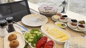 A table with various food items and a bread basket labeled "Mavrella Bozzocoda" on a terrace with a view of buildings and the sea. - Mavrella Bozcaada (Bozcaada)