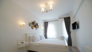 A bedroom with a bed, bedside table, wall-mounted light, decorative wall art, curtains, and a TV. - Mavrella Bozcaada (Bozcaada)