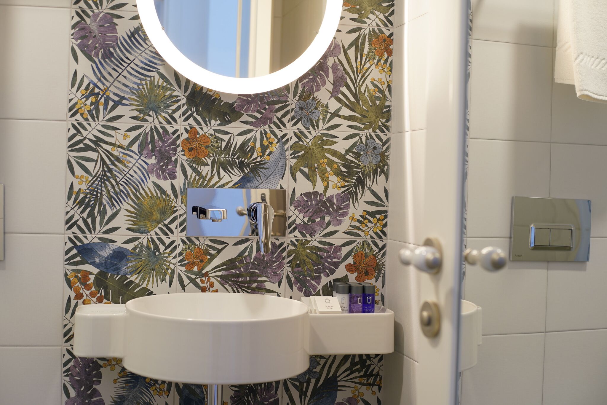 Un baño con salpicadero de azulejos estampados, un espejo redondo, un lavamanos blanco y un perchero para toallas.