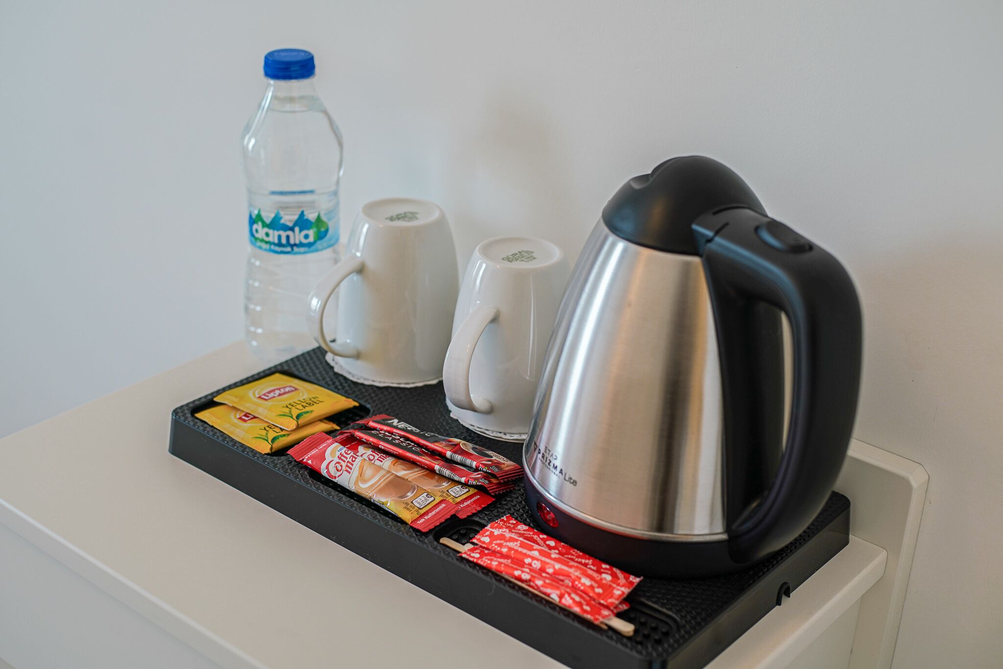 A tray with a bottle of water, two coffee cups, a kettle, and packets of coffee and tea on a white counter.