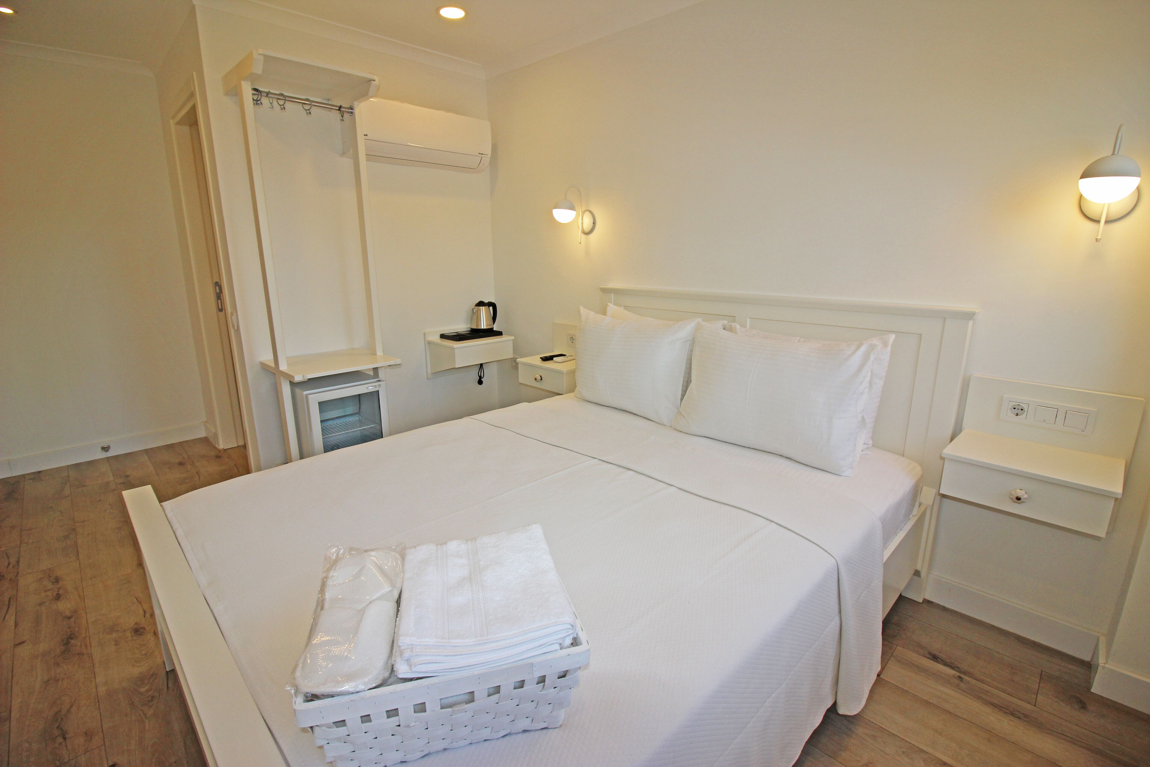 A neatly made bed with white linens, a bedside table with a lamp, and a small shelf with a kettle.