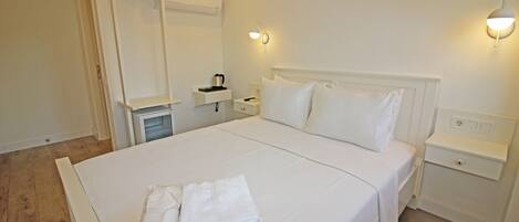 A neatly made bed with white linens, a bedside table with a lamp, and a small shelf with a kettle.