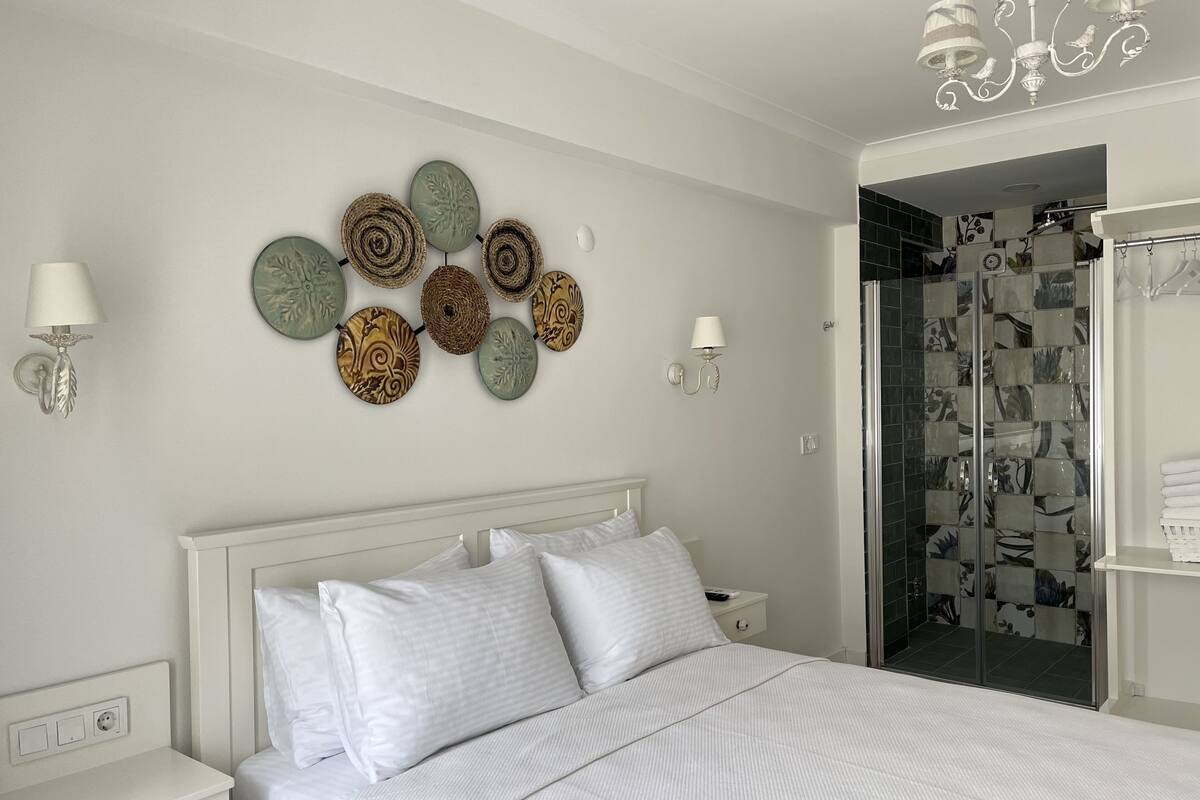 Un dormitorio ordenado con cama, mesitas de noche, lámparas fijadas a la pared y una zona de ducha con azulejos estampados.