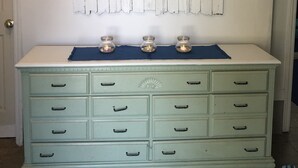 A room with a white wall, a decorative wooden plank, and a green dresser with drawers. - Beach, Please! (Kill Devil Hills)