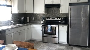 A modern kitchen with white cabinets, a stainless steel refrigerator, a black stove, and a tiled backsplash. - Beach, Please! (Kill Devil Hills)