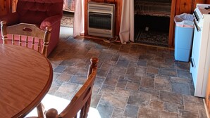 Ein gemütliches Blockhaus-Interieur mit Holzvertäfelung, einem Esstisch mit Stühlen, einem rot gepolsterten Sessel, einem alten Herd und einem kleinen Heizgerät.