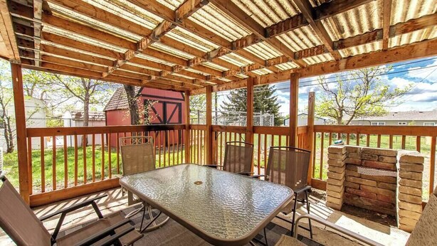 Une terrasse couverte avec une table et des chaises à plateau en verre, une cheminée en briques et une vue sur un bâtiment rouge et des arbres.