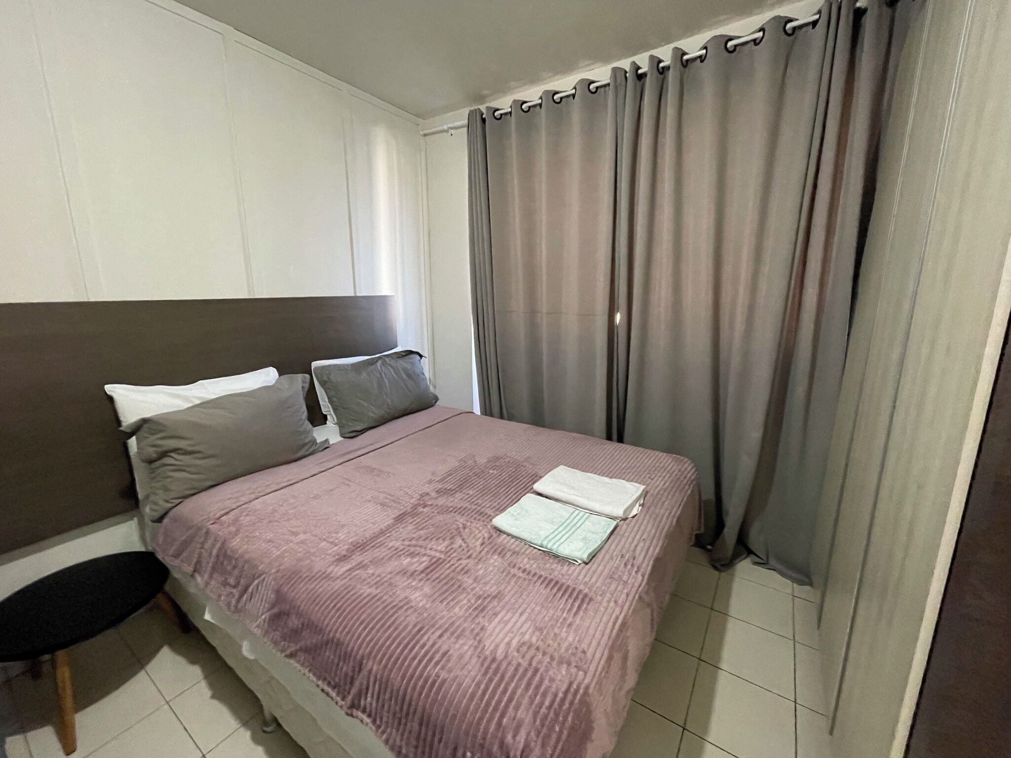 A neatly made bed with a purple bedspread and two pillows, a small table with a chair, and a window with grey curtains.