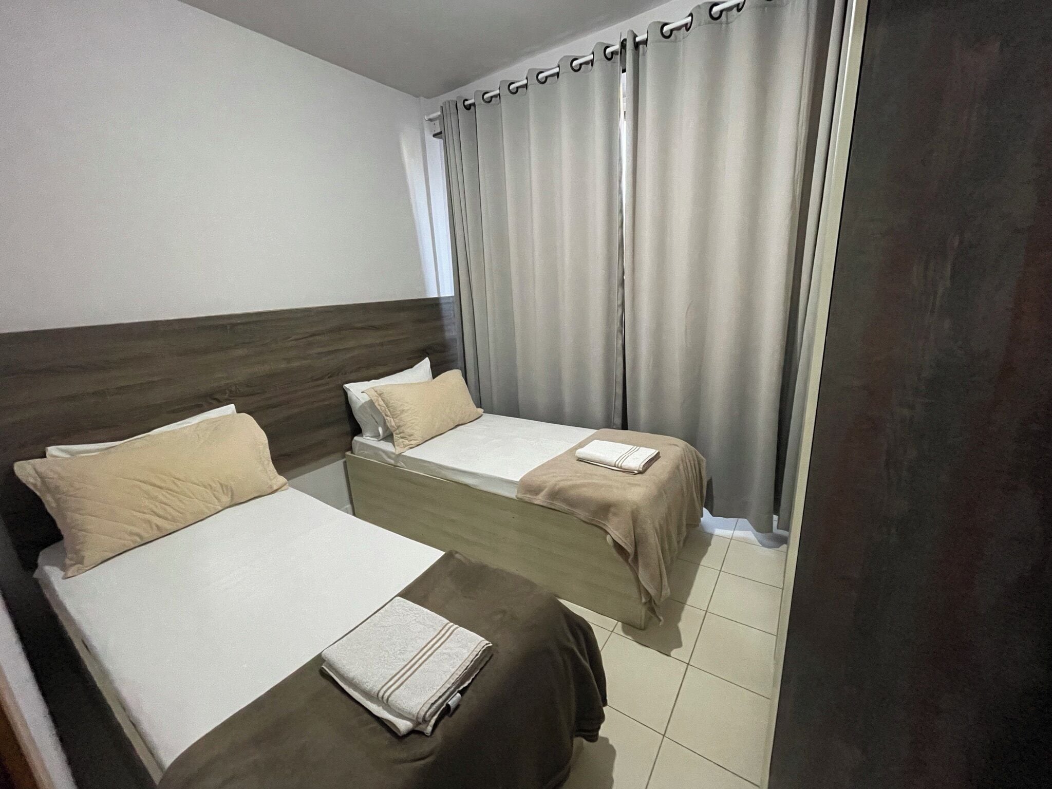 A hotel room with two beds, a wooden headboard, a grey curtain, and a tiled floor.