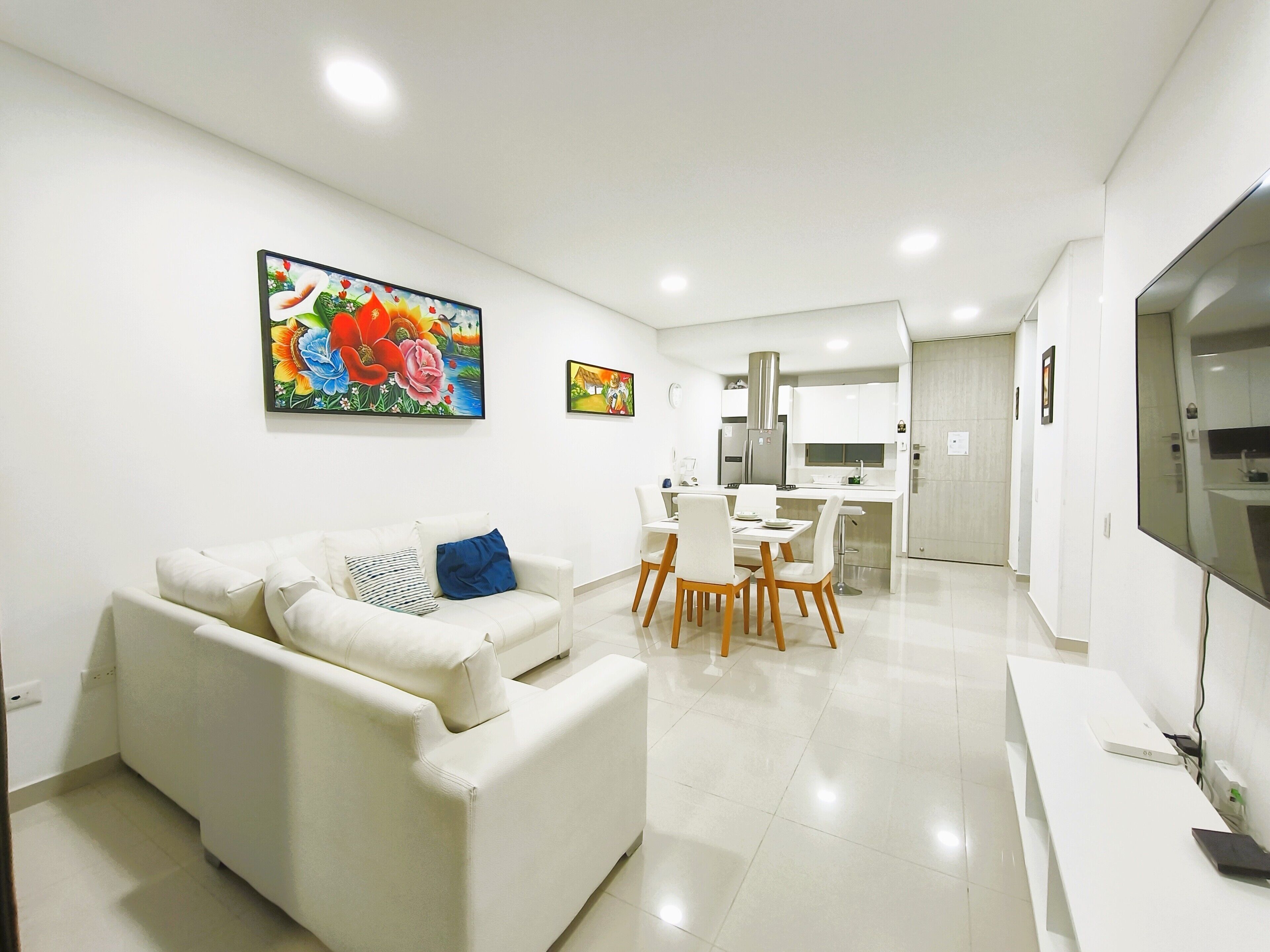 A modern living room with a white sofa, a dining area with a table and chairs, and a kitchen in the background.
