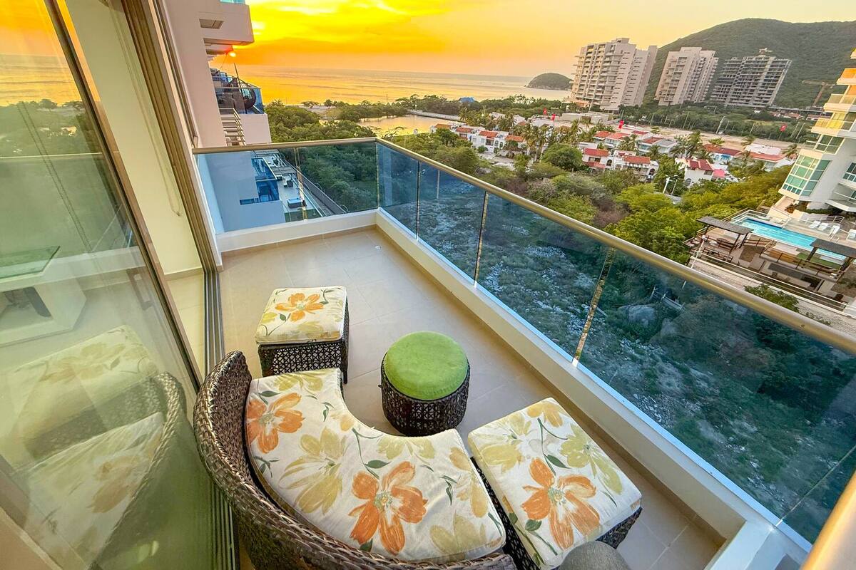 Ein Balkon mit Korbstuhl und Kissen, mit Blick auf eine Stadtlandschaft und ein Gewässer bei Sonnenuntergang.