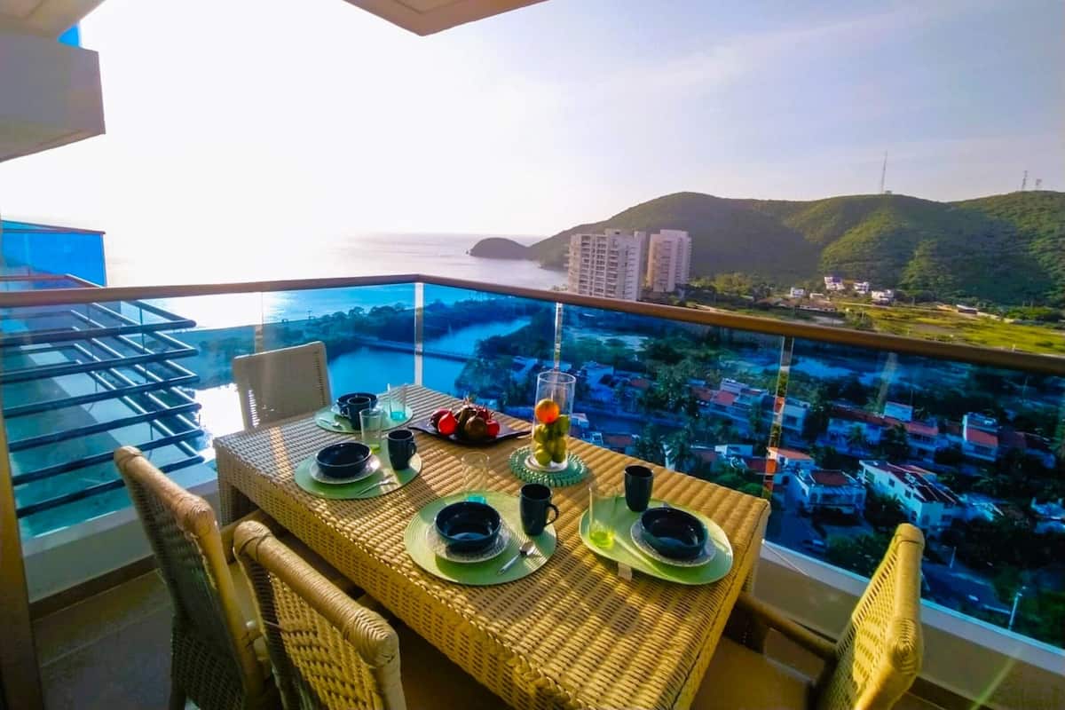 Ein Esstisch mit Tellern, Gläsern und Besteck auf einem Balkon mit Blick auf eine Stadtlandschaft und das Meer.