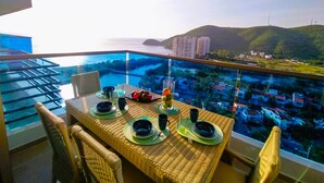 Ein Esstisch mit Tellern, Gläsern und Besteck auf einem Balkon mit Blick auf eine Stadtlandschaft und das Meer.