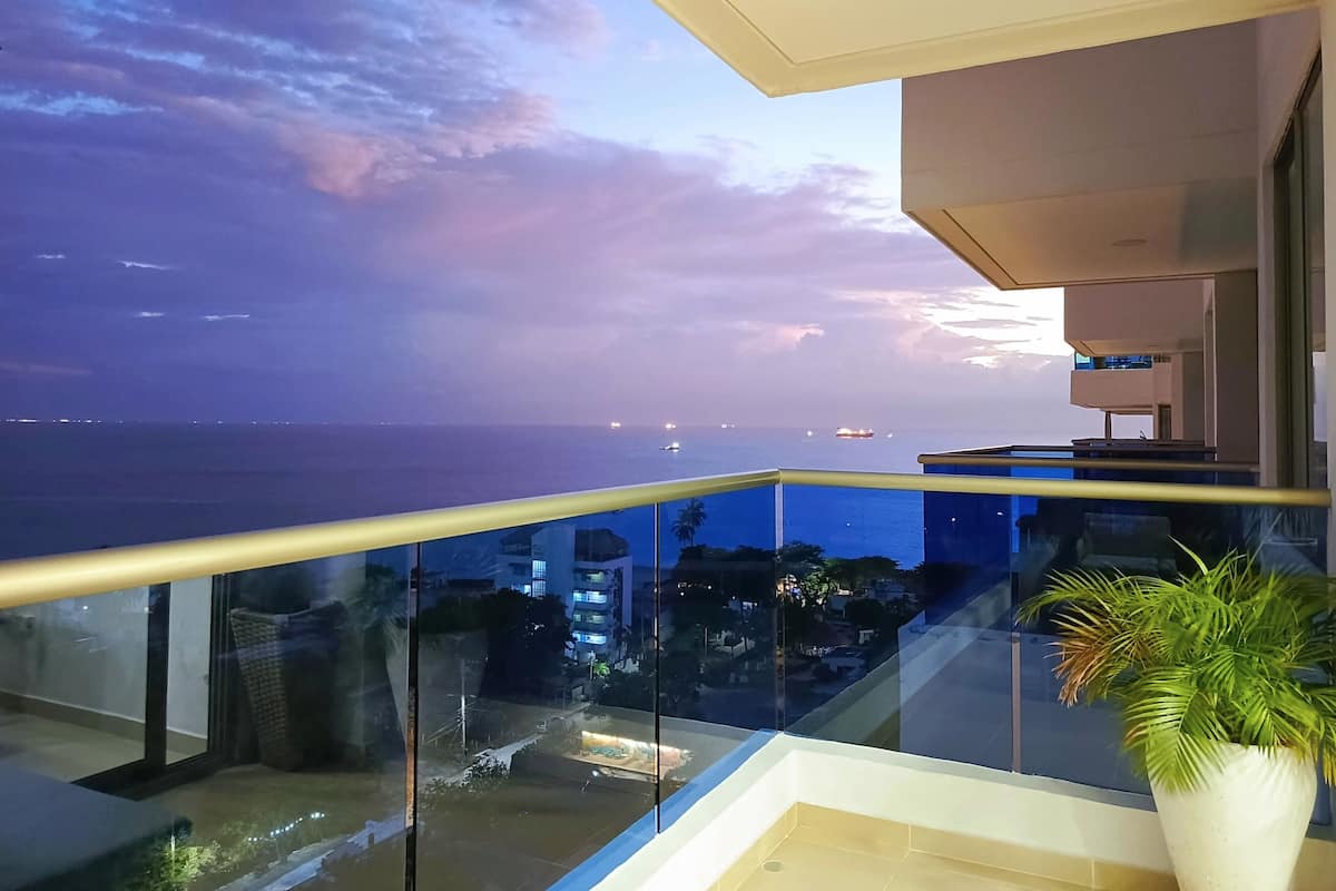Ein Balkon mit Glasgeländer, der auf eine Stadtlandschaft und das Meer im Abendlicht blickt.