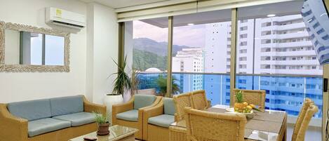 A modern living room with a sofa, chairs, a glass coffee table, and a dining table. There is a large window with a view of buildings and mountains.