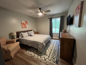 Ein Schlafzimmer mit Bett, Sessel, Nachttisch, Deckenventilator, Fernseher und einem Fenster mit Vorhängen.
