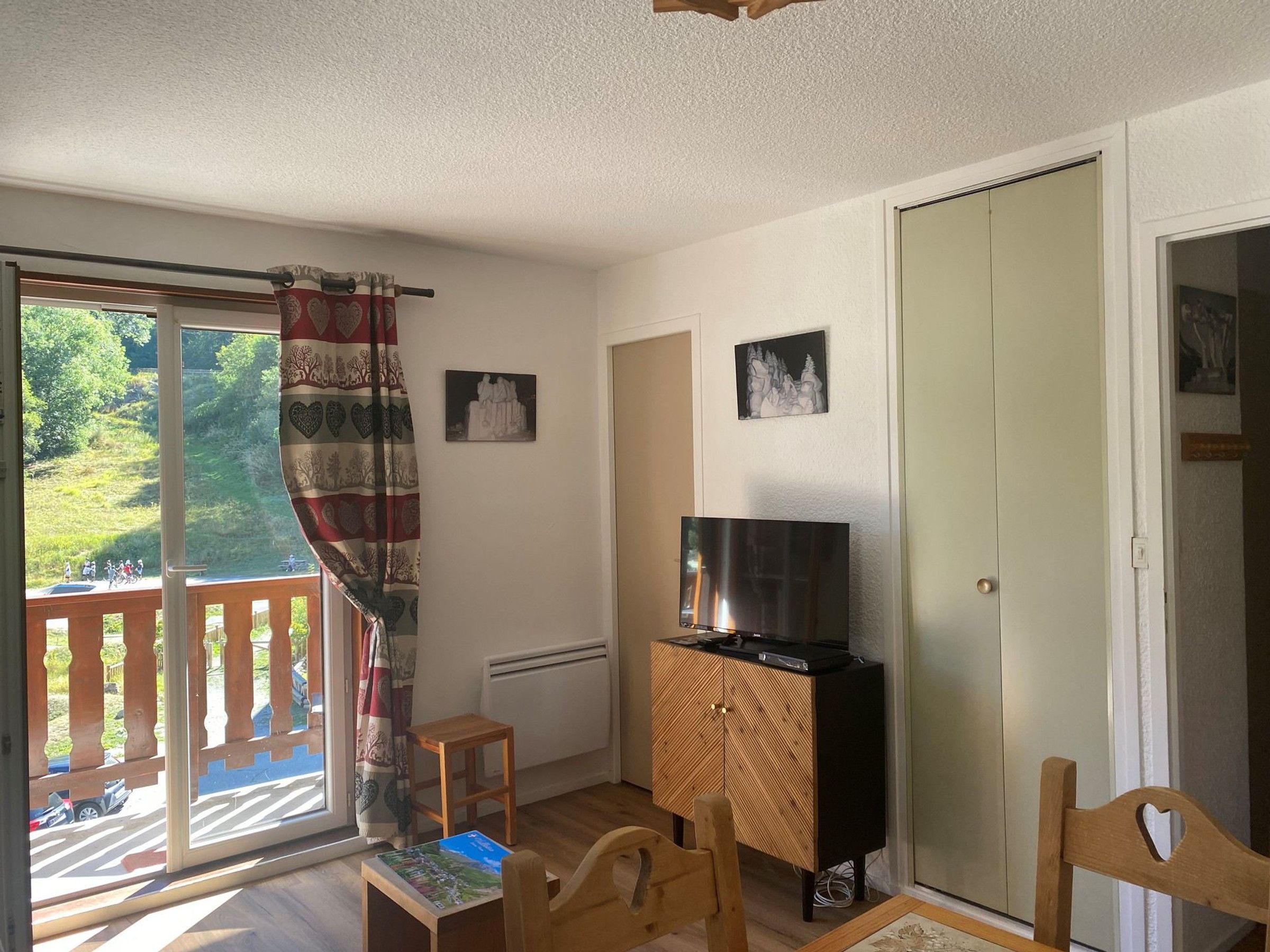 Une pièce comprenant un téléviseur posé sur un meuble en bois, une petite table, une chaise, une fenêtre donnant sur un paysage verdoyant et une porte.