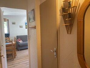 A room with a wooden door, a small table with chairs, a sofa, and a wall-mounted shelf with items.