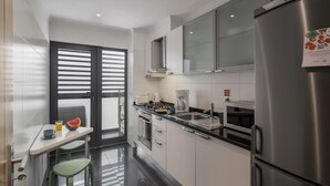 A modern kitchen with a stainless steel refrigerator, a coffee maker, and a microwave. There is a window with blinds, a small dining table with a chair, and a countertop with a sink. - Funchal Holidays, near the beach - Lido View Calla (Funchal)