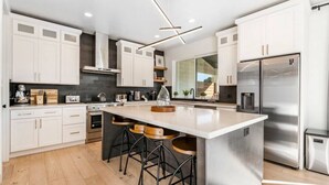 Une cuisine moderne avec des meubles blancs, un grand îlot central doté de tabourets, des appareils en acier inoxydable et un luminaire suspendu au design contemporain.