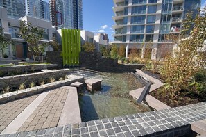 Stroll through a serene oasis amidst the city's modern high-rise buildings, surrounded by lush greenery and a tranquil water feature.