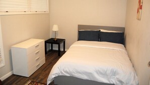 Un dormitorio con cama, cómoda, mesita de noche con lámpara y ventana con persianas.