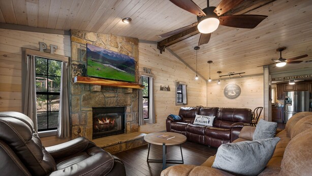 A cozy living room with leather sofas, a stone fireplace, a TV mounted on the wall, and a ceiling fan. - Pet Friendly! Hot Tub | Pool Table | Fire Pit (Pigeon Forge)