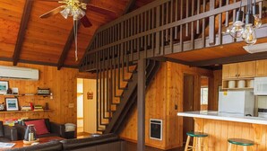 Un intérieur spacieux en bois, avec un plafond haut, un escalier, un coin cuisine et un espace salon comprenant une table et des chaises.
