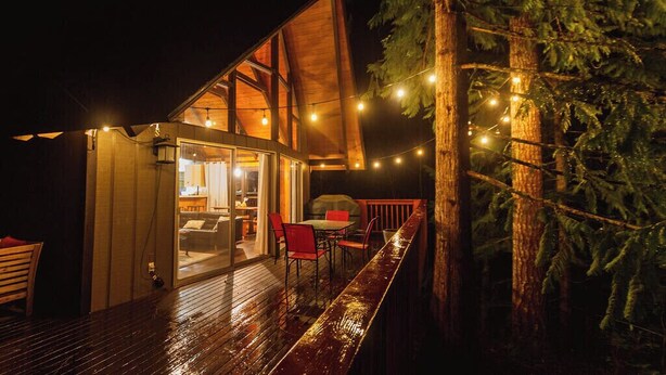 Une terrasse en bois avec un espace repas fermé par des parois de verre, un barbecue et des guirlandes lumineuses.