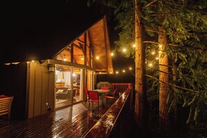 Une terrasse en bois avec un espace repas fermé par des parois de verre, un barbecue et des guirlandes lumineuses.