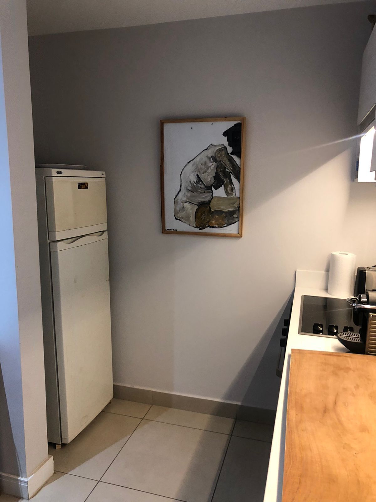 A compact kitchen with a refrigerator, a coffee machine, and a framed artwork on the wall.